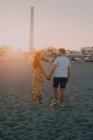 Jovens felizes apaixonados caminhando olhando uns para os outros e de mãos dadas descalços à beira-mar à luz do sol — Fotografia de Stock