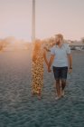 Jóvenes felices enamorados caminando mirándose unos a otros y tomándose de las manos descalzos en la playa a la luz del sol - foto de stock