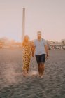 Jovens felizes apaixonados caminhando olhando uns para os outros e de mãos dadas descalços à beira-mar à luz do sol — Fotografia de Stock