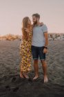 Jóvenes felices enamorados de pie mirándose unos a otros y abrazándose descalzos en la playa a la luz del sol - foto de stock
