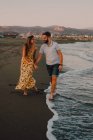 Счастливые влюблённые молодые люди ходят, глядя друг на друга и держась за руки босиком на берегу моря при солнечном свете — стоковое фото