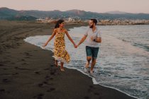 Jóvenes felices enamorados corriendo mirándose los unos a los otros y tomándose de las manos descalzos en la playa a la luz del sol - foto de stock