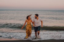 Jóvenes felices enamorados corriendo mirándose los unos a los otros y tomándose de las manos descalzos en la playa a la luz del sol - foto de stock