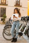 Joyeuse jeune femme en tenue décontractée assis sur un vélo de location sur la station de partage et regardant la caméra sur la rue de la ville — Photo de stock
