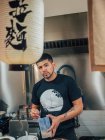 Молодой этнический мужчина готовит японское блюдо под названием рамен и смотрит в камеру в азиатском кафе — стоковое фото