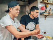 Side view on Asian man offering sauce for fresh cooked Japanese dish ramen and ethnic colleague standing nearby — Stock Photo