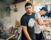 Ethnischer Mann steht und schaut in die Kamera, während Kollege Schüsseln mit Ramen in der Küche serviert — Stockfoto