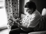Vue latérale de la femme âgée en chemisier et lunettes assis sur un fauteuil près de la fenêtre et la résolution de mots croisés à la maison — Photo de stock