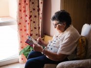 Vista lateral da mulher idosa em blusa e óculos sentados na poltrona perto da janela e resolvendo palavras cruzadas em casa — Fotografia de Stock