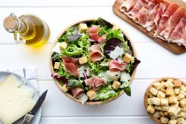 De arriba tazón de deliciosa ensalada de lechuga con tocino y croutons colocados en la mesa blanca cerca de la botella de aceite y rebanada de queso - foto de stock