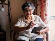 Старша жінка в сорочці і окулярах сидить на кріслі і читає електронну книгу в квартирі — стокове фото