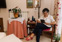 Seniorin in Hemd und Brille sitzt auf Sessel und liest E-Book in Wohnung — Stockfoto