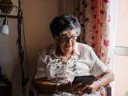 Mujer mayor en camisa y gafas sentada en sillón y leyendo e-book en apartamento - foto de stock