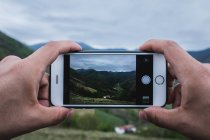 Cerrar las manos de viajeros anónimos usando smartphone para tomar fotos de terrenos verdes montañosos y pueblos en días nublados. - foto de stock