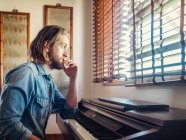 Thoughtful young man looking through window near synthesizer at home — Stock Photo