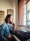 Продуманий молодий чоловік дивиться крізь вікно біля синтезатора вдома — стокове фото