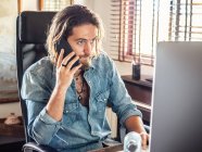 Konzentrierter Mann im lässigen Hemd kommuniziert auf dem Smartphone, während er den Computer im Home Office benutzt — Stockfoto