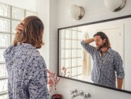 Прохолодний молодий чоловік фіксує волосся стоячи у світлій ванній перед дзеркалом — стокове фото