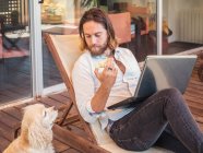 Homme avec ordinateur portable et noyau de pomme avec chien d'épagneul demandant sur le balcon — Photo de stock