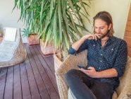 Модний красивий молодий чоловік сидить у кріслі з телефоном на терасі — стокове фото