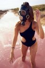 Woman in swimsuit and respirator mask walking in pink water polluted with plastic bottles at red lagoon — Stock Photo