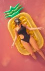 From above woman in respirator mask and swimwear chilling on inflatable mattress in form of pineapple in red lagoon water — Stock Photo