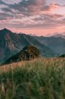 Теплый пейзаж с одиноким камнем в холмистой местности, рядом скалистые горы под красно-серым облачным небом при закатном свете — стоковое фото