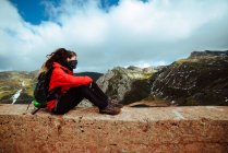 Frau in roter Jacke und Schal sitzt in der Nähe der Landstraße am Berghang an bewölkten Tagen in herrlicher Natur — Stockfoto