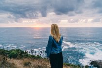 Visão traseira da mulher loira arrepiante e contemplando paisagens cênicas enquanto está sozinha na costa calma em nuvens — Fotografia de Stock