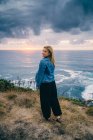 Visão traseira da mulher loira arrepiante e contemplando paisagens cênicas enquanto está sozinha na costa calma em nuvens — Fotografia de Stock