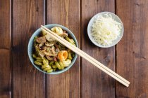 Pastas ecológicas con verduras en tazón y palillos de sushi en escritorio de madera con plato auxiliar - foto de stock