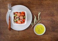De dessus composition de salade avec tomates cerises et olives dans un bol et de l'huile sur le côté servi sur fond de bois minable — Photo de stock