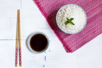 Ansicht der Schüssel mit traditionellem japanischen Reis auf rosa Handtuch und Essstäbchen per Untertasse mit Sojasauce auf weißem Tisch. — Stockfoto