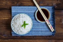 Vista superior del tazón de arroz tradicional japonés en toalla azul y palillos en platillo con salsa de soja en mesa de madera
. - foto de stock