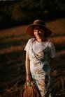 Femme en robe rétro et chapeau marchant dans le champ vers le ciel du coucher du soleil en regardant la caméra — Photo de stock