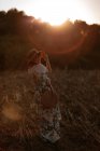 Back view of woman in retro dress and hat walking in field towards sunset sky while looking away — Stock Photo