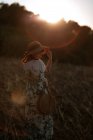 Back view of woman in retro dress and hat walking in field towards sunset sky while looking down — Stock Photo