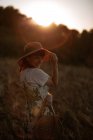 Vista lateral de la mujer con vestido retro y sombrero caminando en el campo hacia el cielo de puesta de sol mientras se mira la cámara. - foto de stock