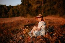 Vista lateral da mulher em vestido retro e chapéu sentado no meio de um campo enquanto olha para a câmera — Fotografia de Stock