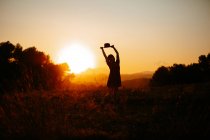 Silhouette of woman raising hands with hat and dancing against bright sunset sky in field — Stock Photo