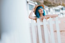 Romantique femme heureuse avec les cheveux bleus teints en chapeau de soleil et robe reposant tout en se penchant dans la clôture à la ville côtière rurale — Photo de stock