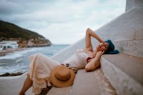 Side view of dreamy woman with colorful blue hair in sundress lying on stone steps on seaside — Stock Photo