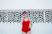 Модная женщина в ярких солнцезащитных очках с синей прической в красном купальнике, стоящая с протянутой рукой рядом с дизайнерской стеной — стоковое фото