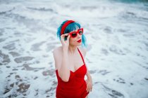 Femme à la mode avec des cheveux bleus en maillot de bain rouge brillant touchant lunettes de soleil rouges dans l'eau mousseuse — Photo de stock