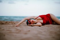 Модна жінка з синіми волосками в червоному яскравому купальнику, яка лежить на піщаному пляжі з розпростертими руками. — стокове фото