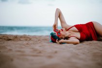 Modische Frau mit blauen Haaren im roten hellen Badeanzug genießt es, am Sandstrand zu liegen — Stockfoto