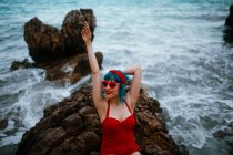 Mulher elegante com cabelos azuis em maiô vermelho brilhante tendo descanso confortavelmente sentado em pedra rochosa escura em água do mar espumosa — Fotografia de Stock