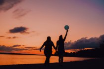 Вид сзади анонимных женщин с воздушными шарами, держащимися за руки и идущими вдоль берега во время красивого заката — стоковое фото