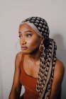 Schöne erwachsene Afroamerikanerin in braunem Kleid mit gemustertem Kopftuch und Ohrringen, die vor grauem Hintergrund wegschauen — Stockfoto