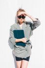 Бізнес-леді з модною зачіскою, сонцезахисними окулярами і костюмом тримає ноутбук і дивиться на камеру на стіні — стокове фото